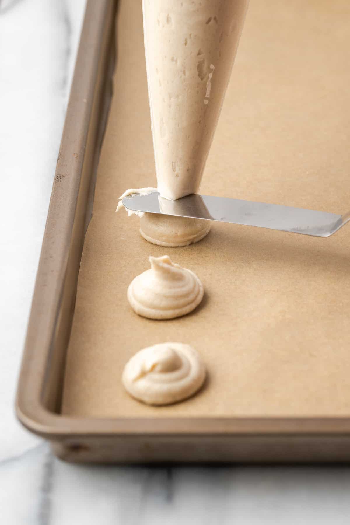 An knife is used to cut the vanilla wafer batter from the end of a piping bag as it's piped onto a lined baking sheet.