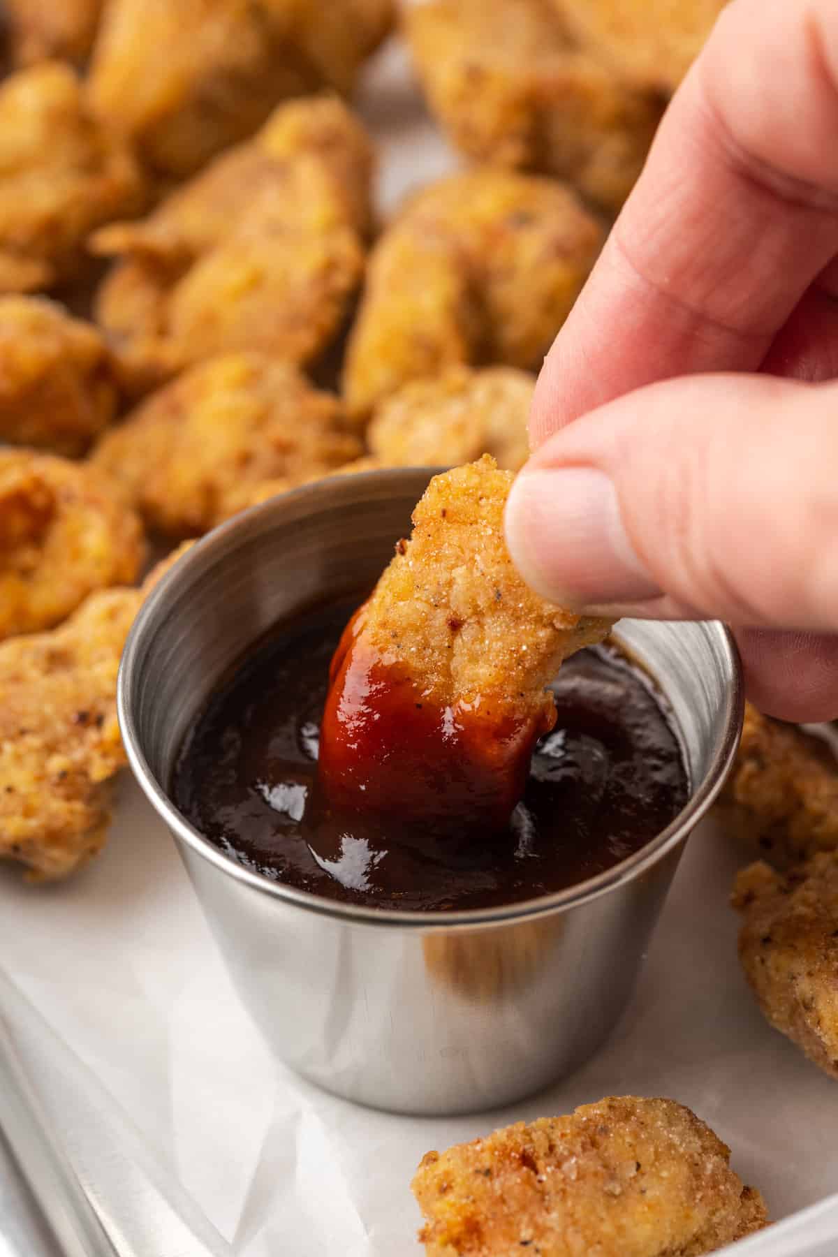 A hand dips a piece of gluten-free popcorn chicken into a ramekin of BBQ sauce.