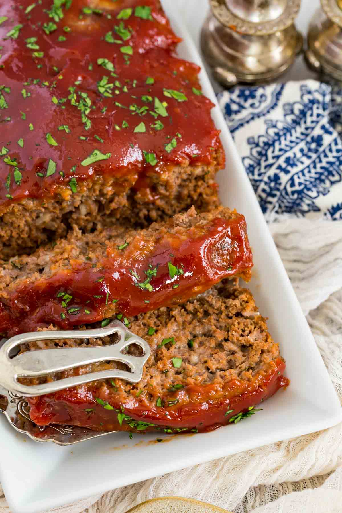 Tongs are used to lift a slice of gluten-free meatloaf from a plate.