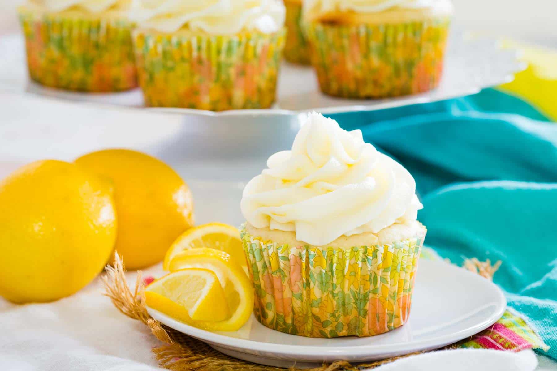 A frosted gluten-free lemon cupcake on a white plate next to lemon slices, with more cupcakes on a cupcake stand in the background.