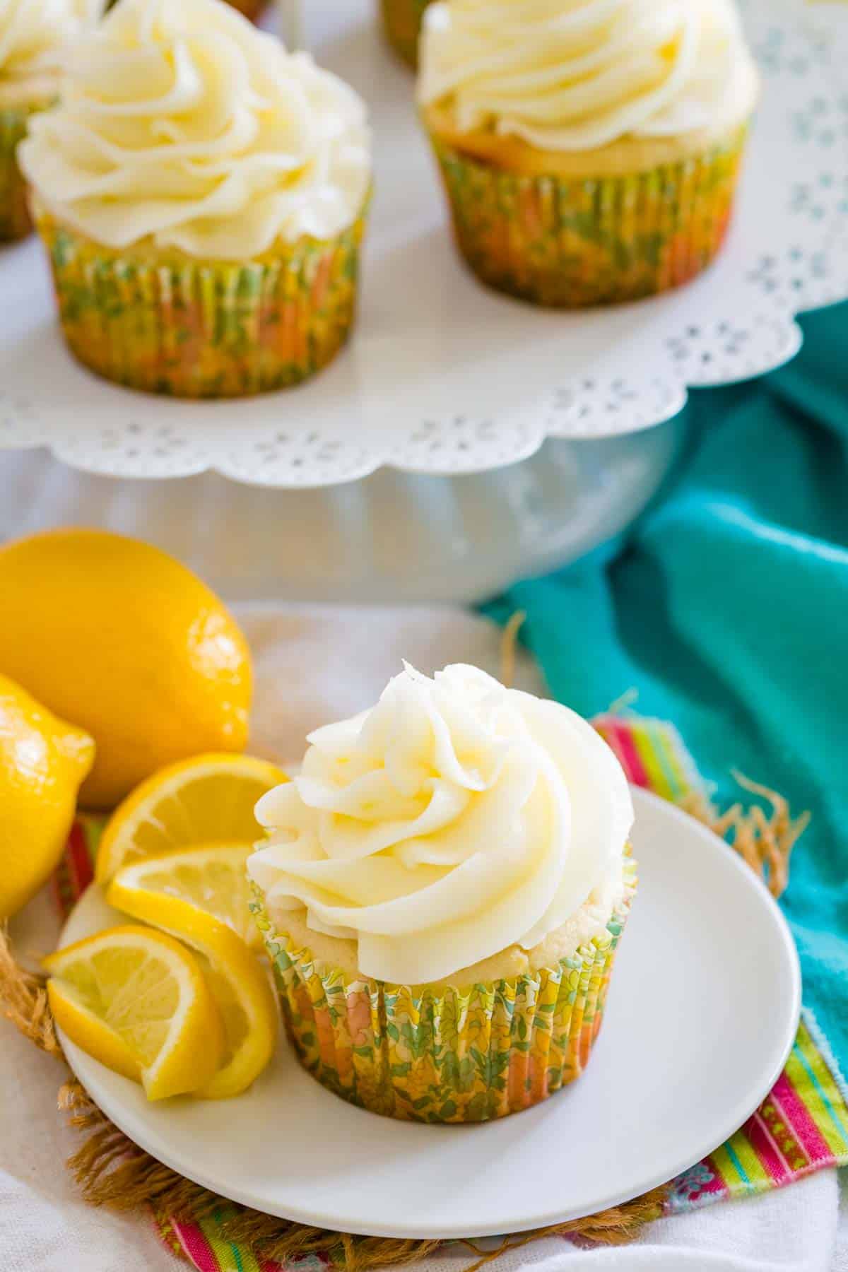 A frosted gluten-free lemon cupcake on a white plate next to lemon slices, with more cupcakes on a cupcake stand in the background.