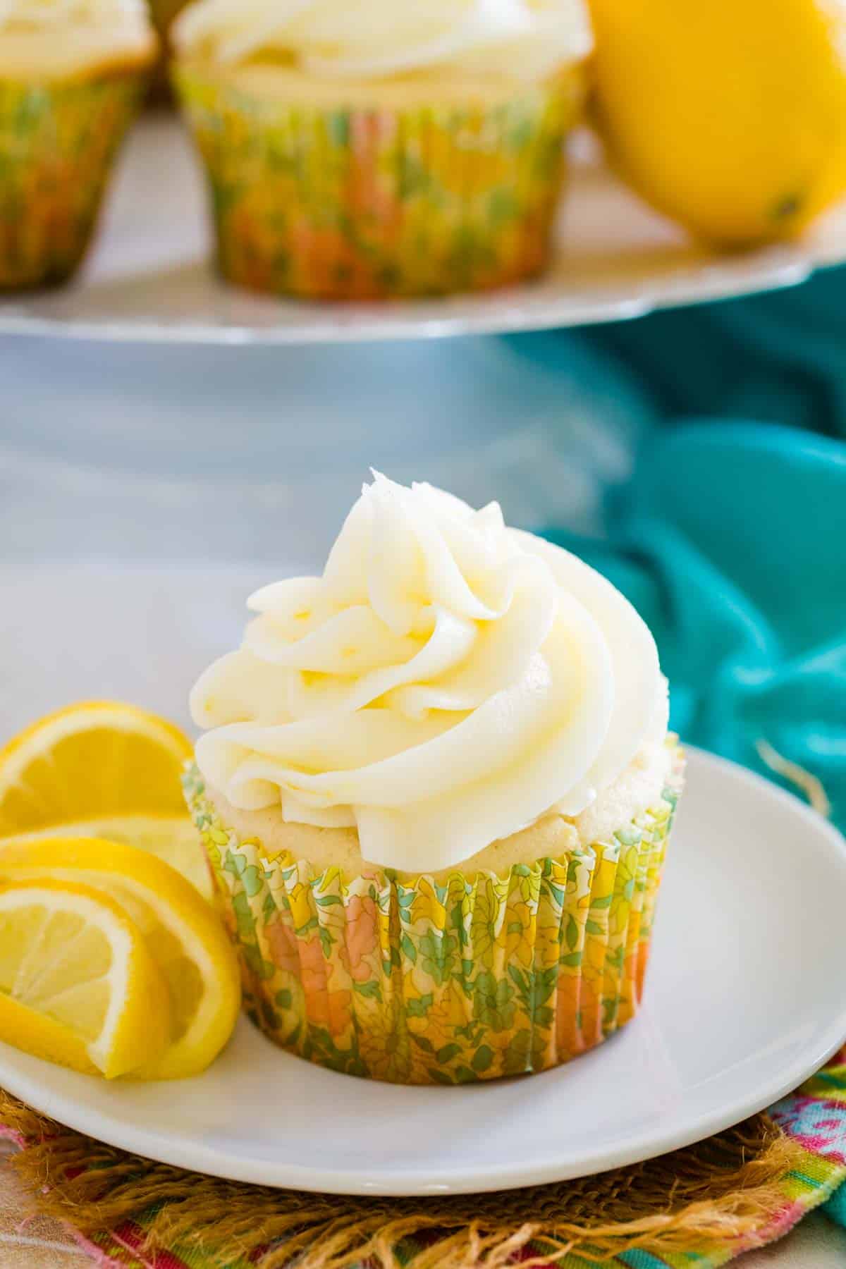 A frosted gluten-free lemon cupcake on a white plate next to lemon slices, with more cupcakes on a cupcake stand in the background.