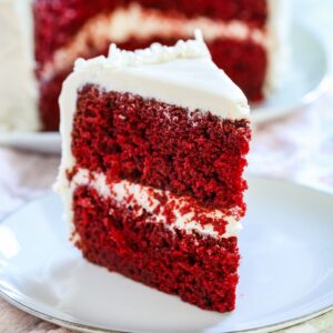 A piece of cake with tow layers of red velvet with cream cheese frosting between them and on top on a small white plate.