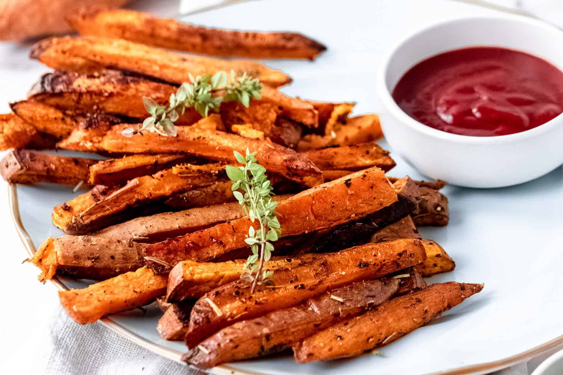 Herbes de Provence baked sweet potato fries on a white plate next to a bowl of ketchup.