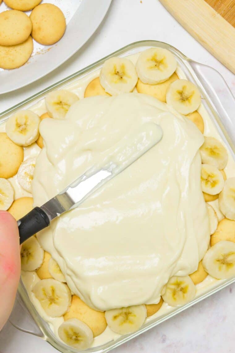 A hand uses an offset spatula to spread a pudding layer over top of the banana layer inside a casserole dish of banana pudding.