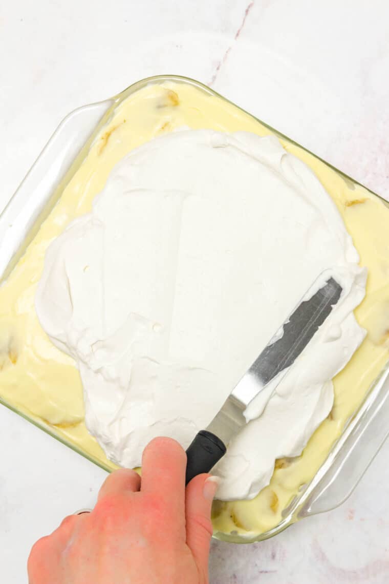 A hand uses an offset spatula to spread a final layer of whipped cream over a casserole dish of chilled banana pudding.