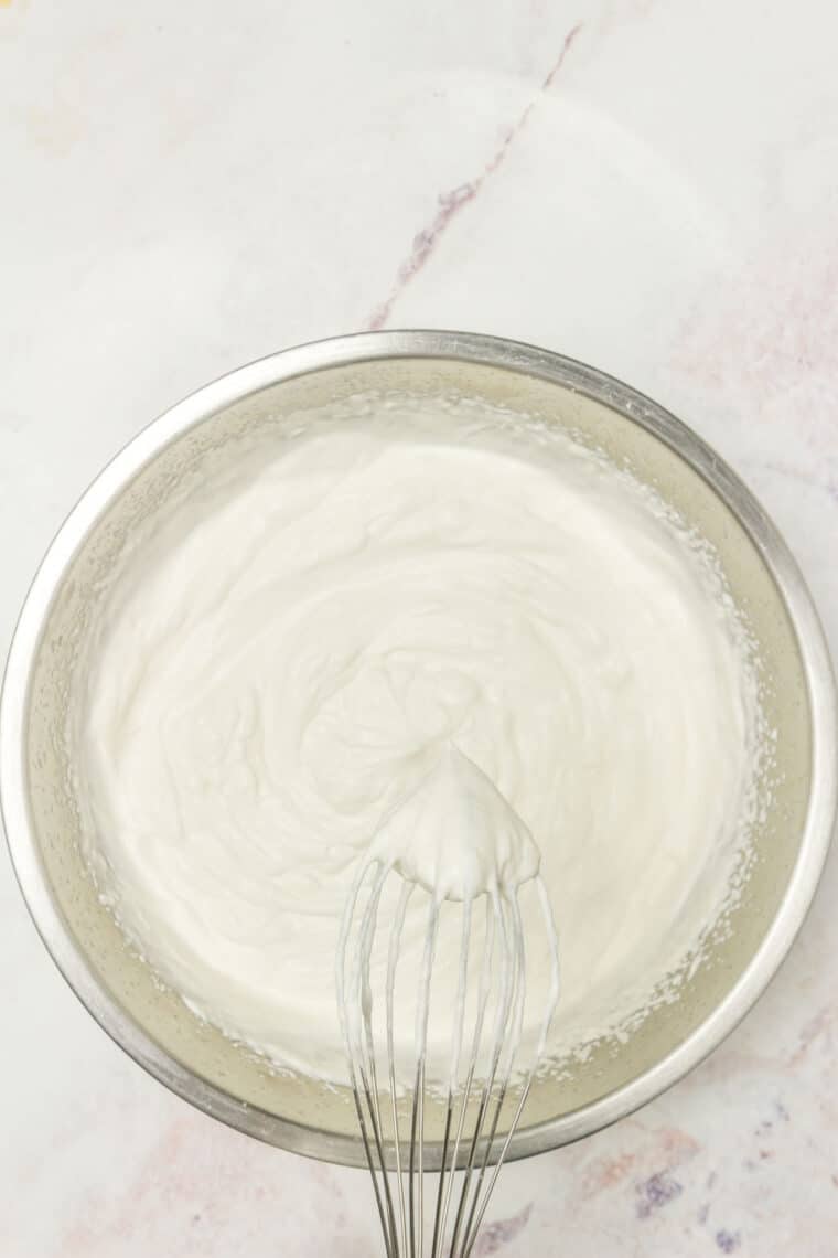 A whisk with a stiff peak of whipped cream on the tip held above a bowl of freshly whipped cream.