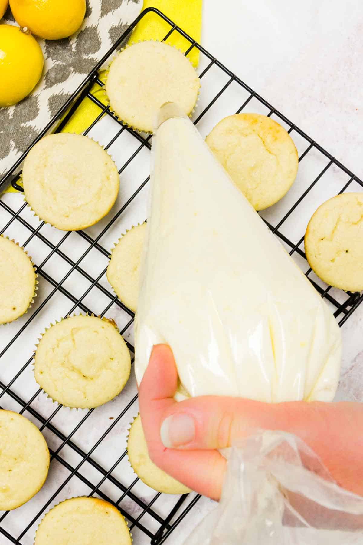 A hand holds a piping bag filled with lemon frosting over rows of baked gluten-free lemon cupcakes on a wire rack.