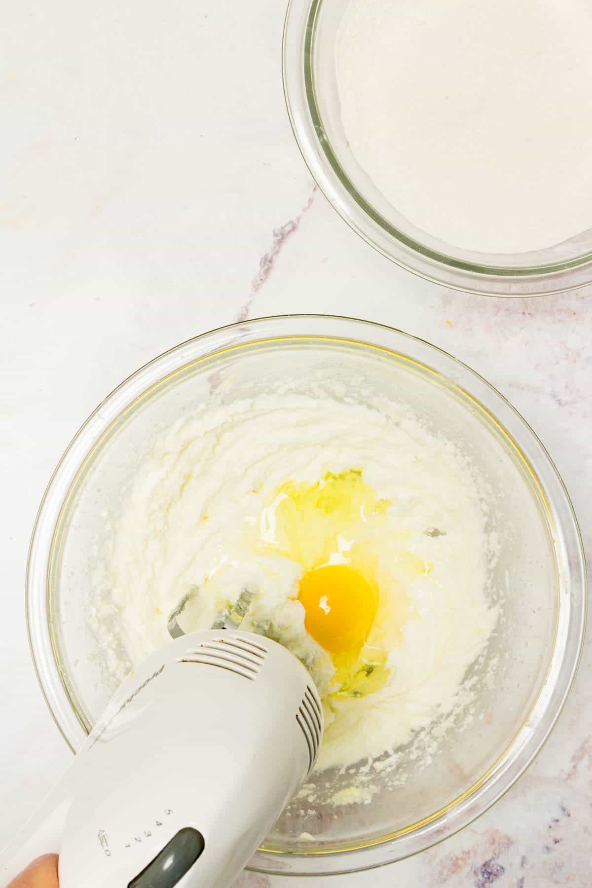 A hand blender mixes egg into a mixing bowl with creamed butter and sugar.