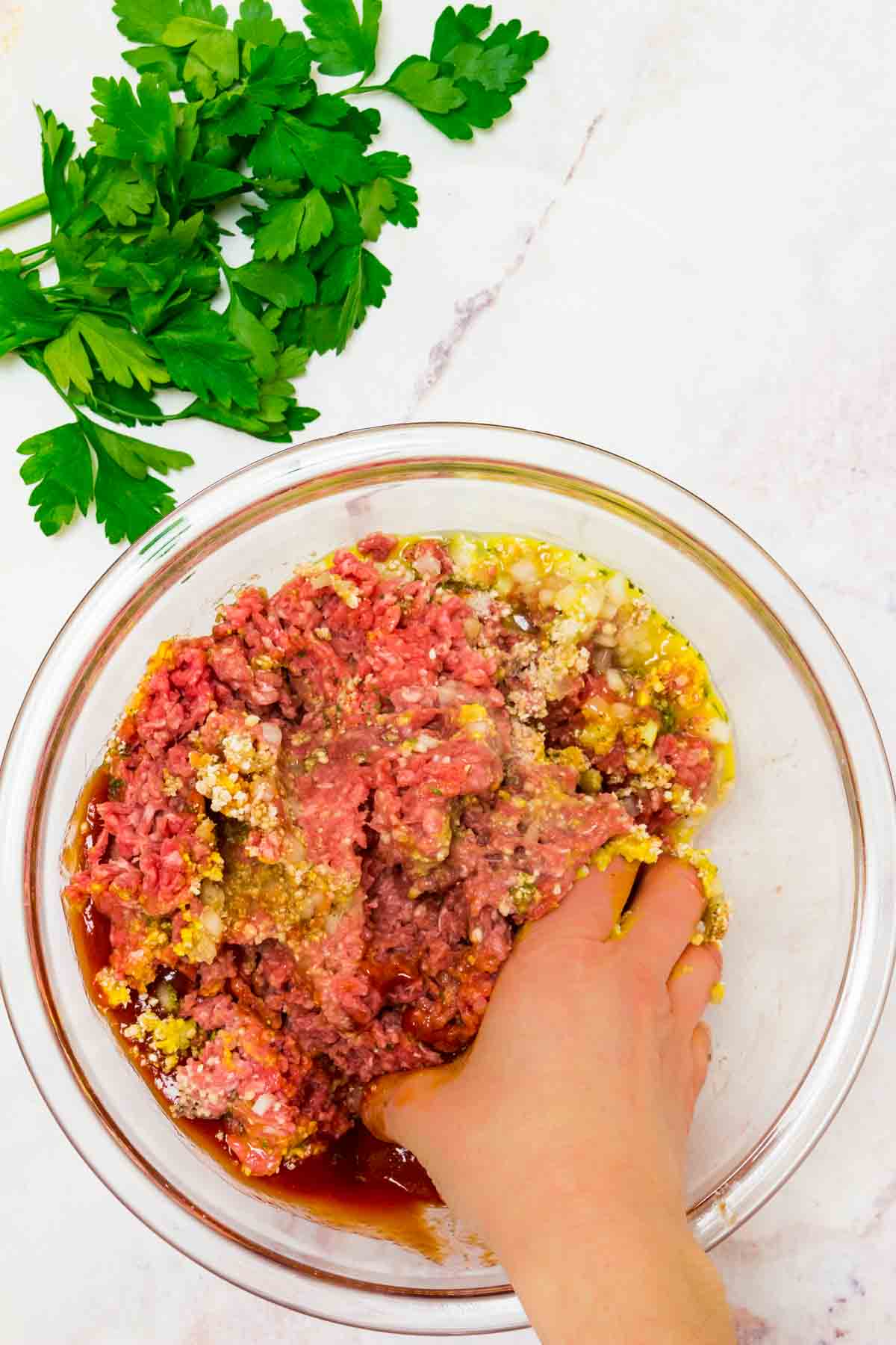 A hand mixing ground beef and meatloaf ingredients inside a glass bowl.