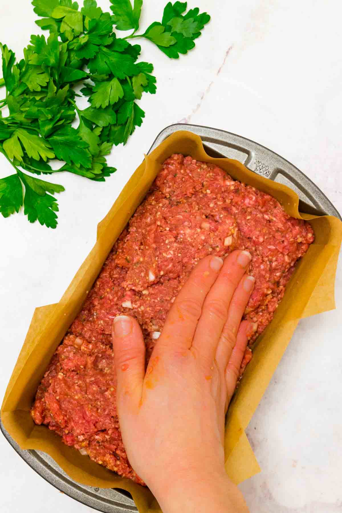 A hand presses the meatloaf mixture into a rectangular baking pan lined with parchment paper.