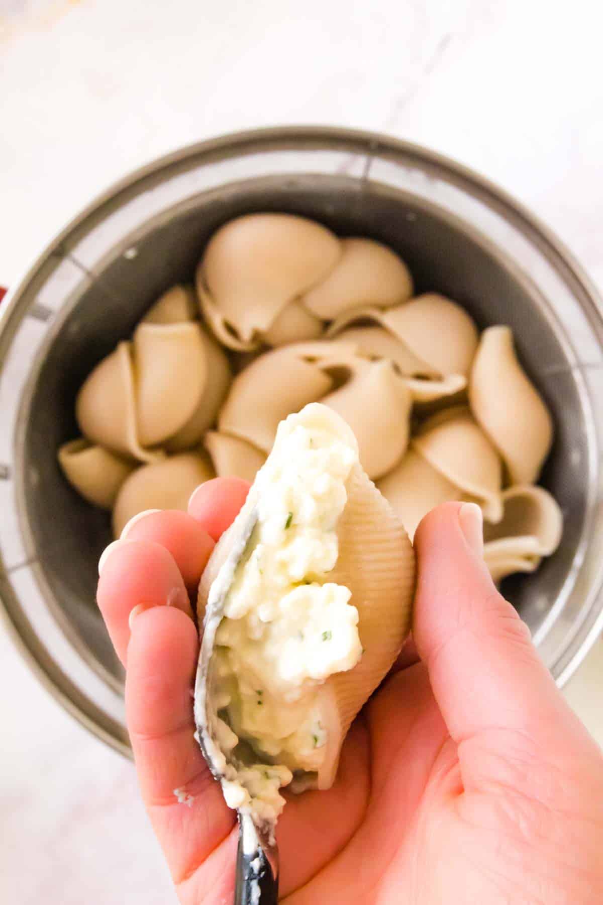 Overhead view of a hand filling a pasta shell with cheese, with a bowl of gluten-free jumbo pasta shells below.