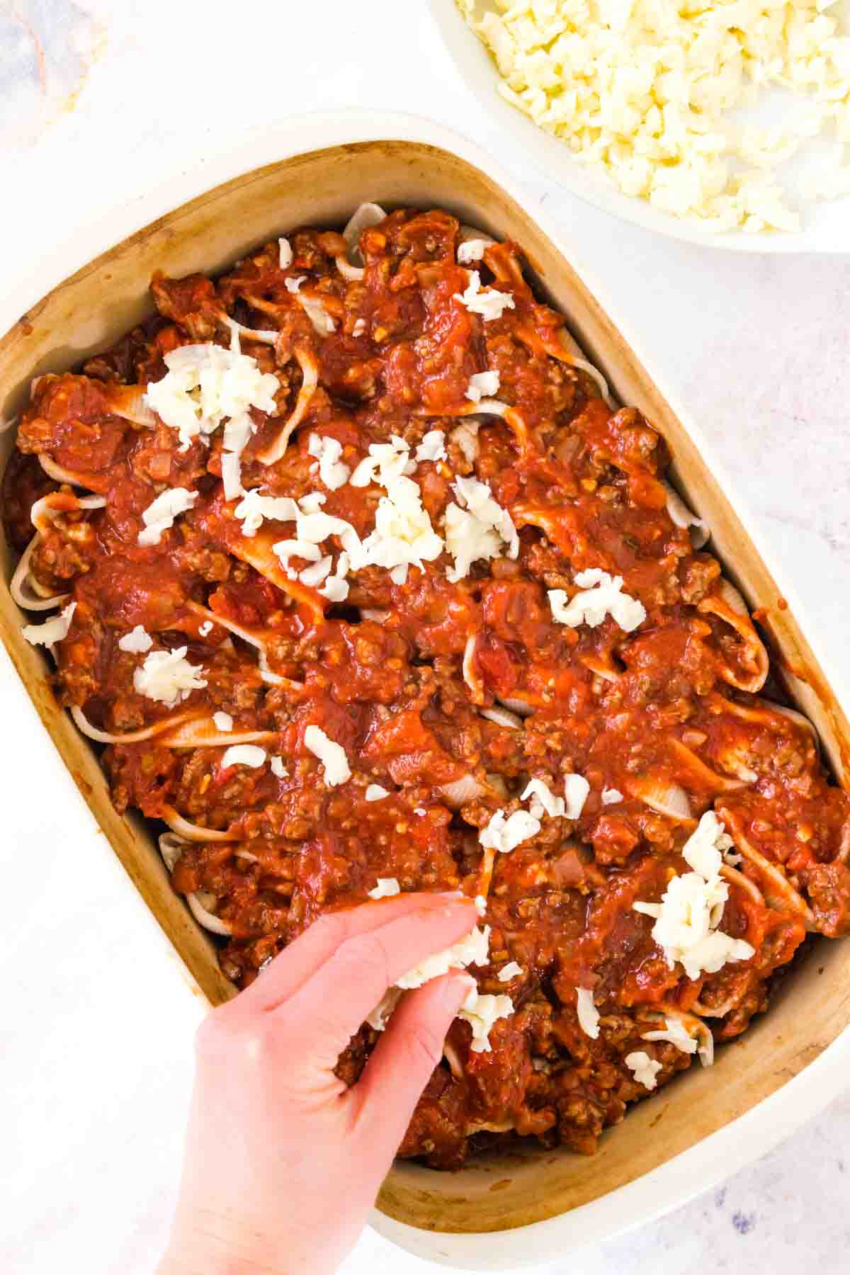 A hand sprinkles mozzarella cheese over layers of gluten-free stuffed shells and meat sauce in a casserole dish.