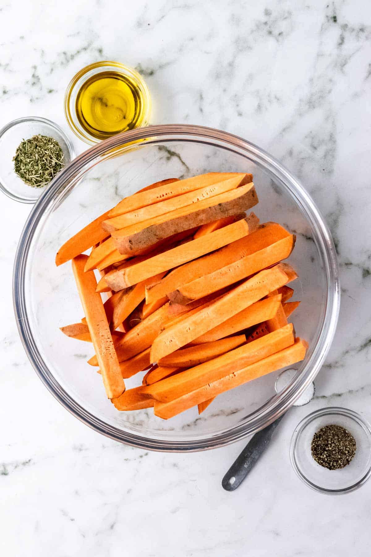 Cut sweet potatoes in a glass bowl next to bowls of oil and seasonings.