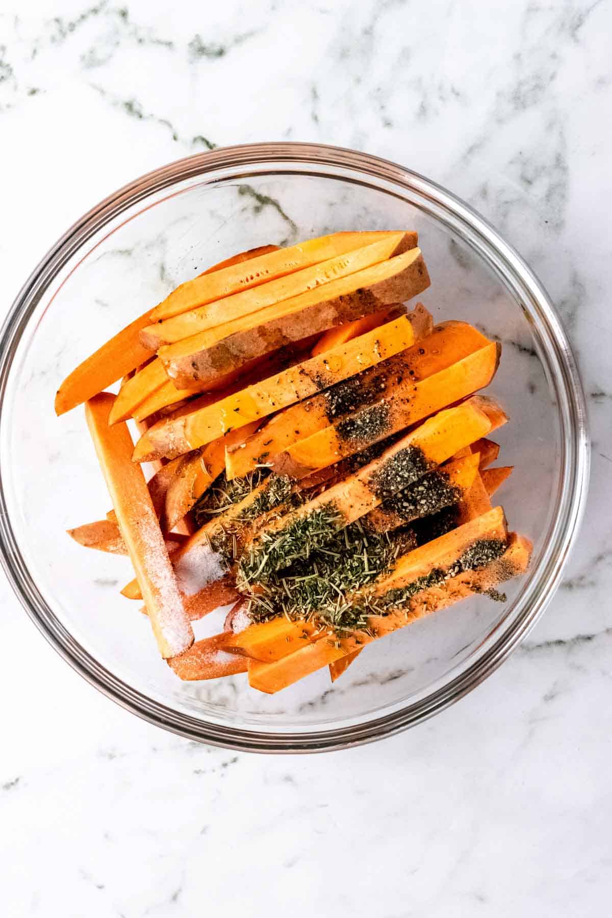 Cut sweet potatoes combined with Herbes de Provence seasoning in a glass bowl.