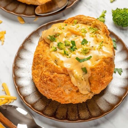 A bread bowl on a brown scallop-edge plate overflowing with broccoli cheese soup.