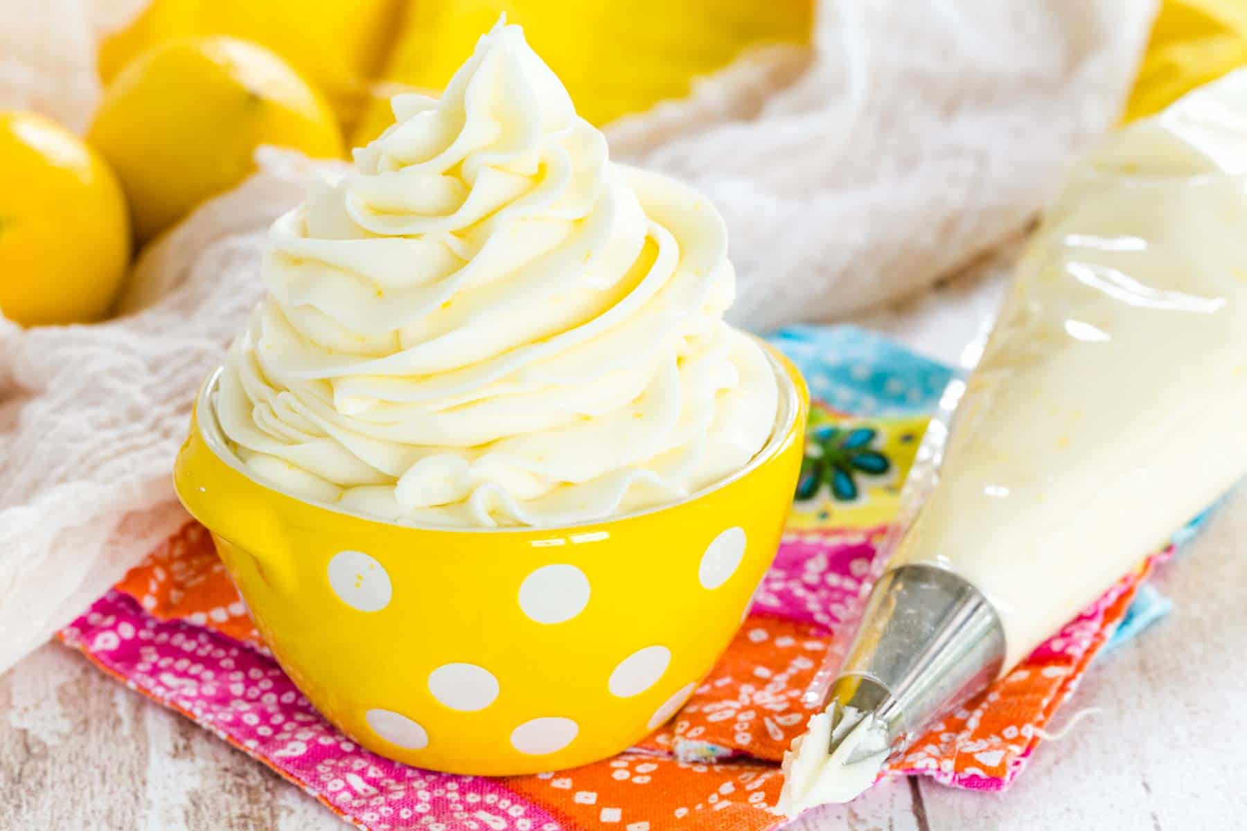 A swirl of lemon buttercream frosting in a small yellow bowl with white polka dots, next to a piping bag with lemons in the background.