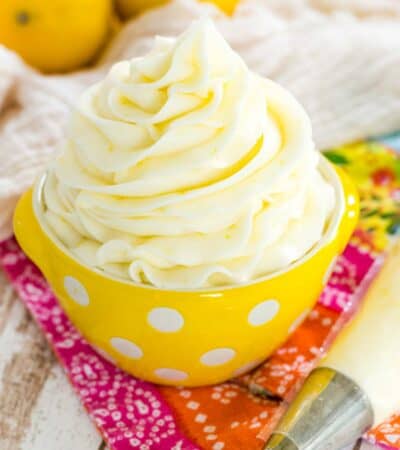 A swirl of lemon buttercream frosting in a small yellow bowl with white polka dots, next to a piping bag with lemons in the background.