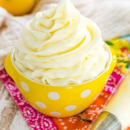 A swirl of lemon buttercream frosting in a small yellow bowl with white polka dots, next to a piping bag with lemons in the background.