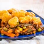 A serving of ground beef and veggies topped with tater tots and cheese on a blue plate with text overlay that says "Tater Tot Shepherd's Pie".