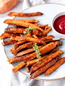 Herbes de Provence baked sweet potato fries on a white plate next to a bowl of ketchup.