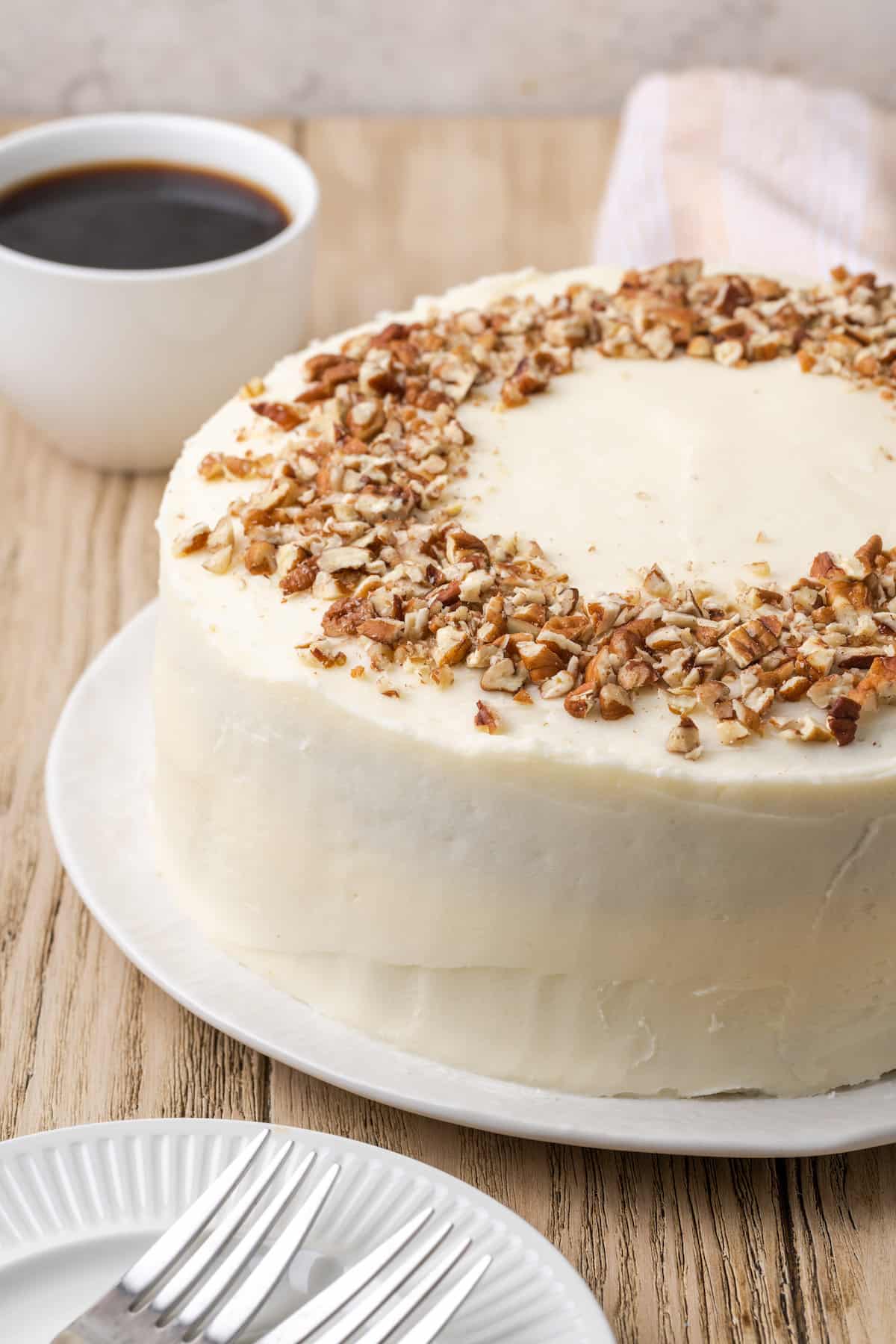 Frosted gluten-free carrot cake on a white plate, topped with a ring of crushed pecans with a cup of coffee in the background.