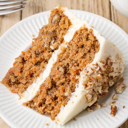 A slice of frosted gluten-free carrot cake on a white plate next to a fork and a cup of coffee.