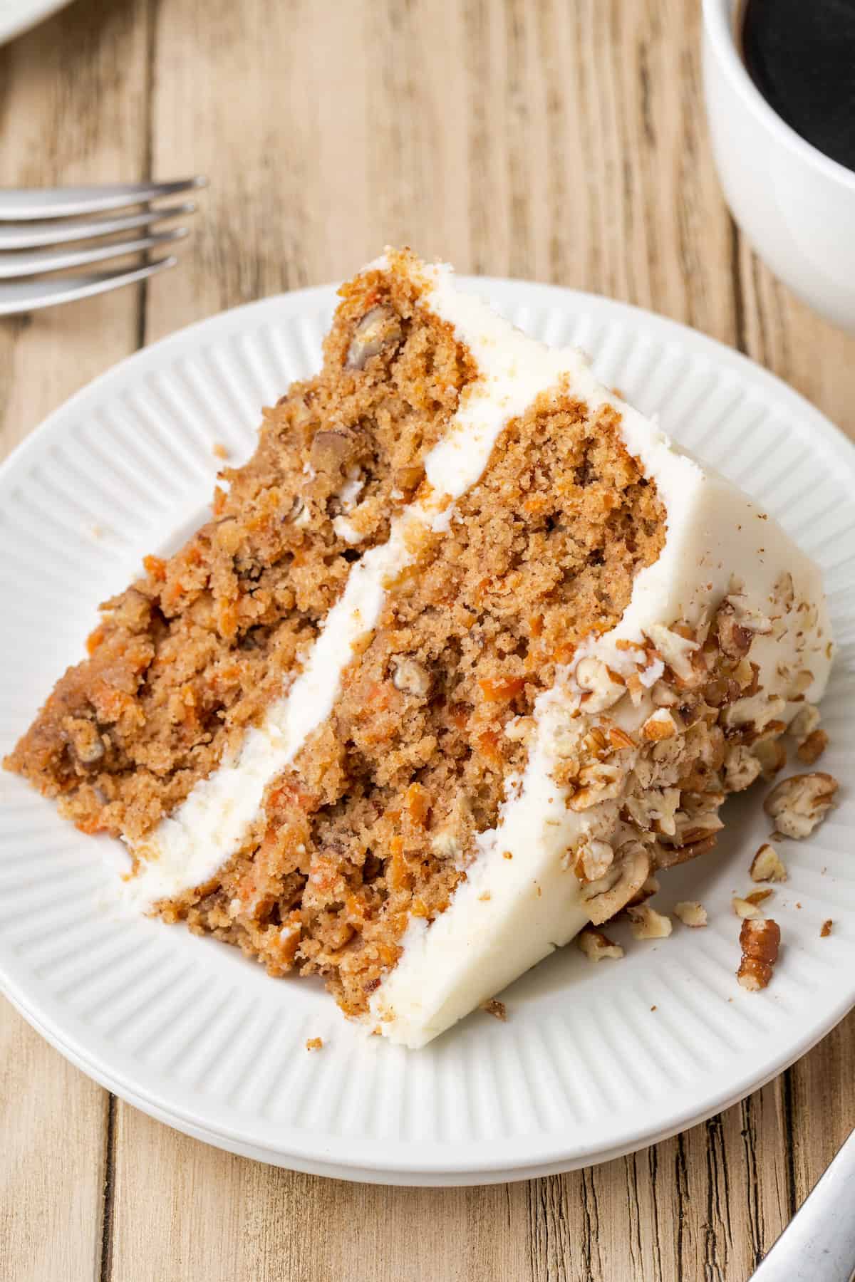 A slice of frosted gluten-free carrot cake on a white plate next to a fork and a cup of coffee.