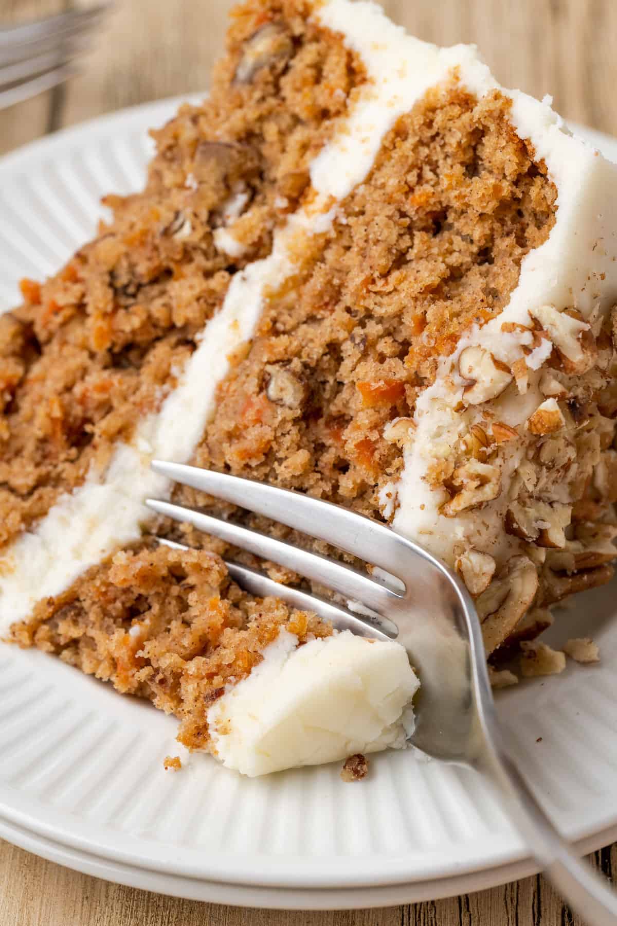 A fork cuts into the corner of a slice of frosted gluten-free carrot cake on a white plate.