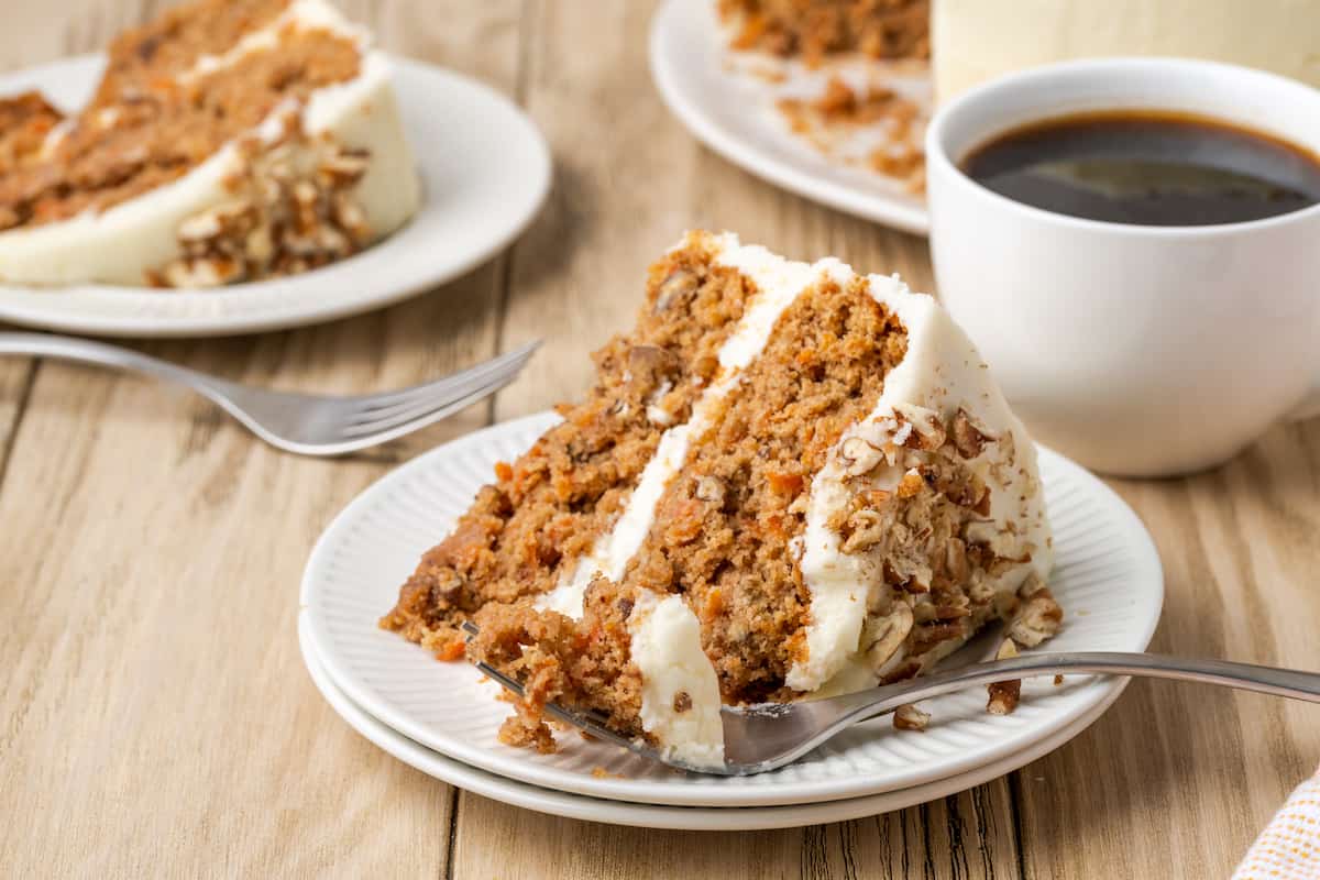 A slice of frosted gluten-free carrot cake on a white plate with a cup of coffee in the background.