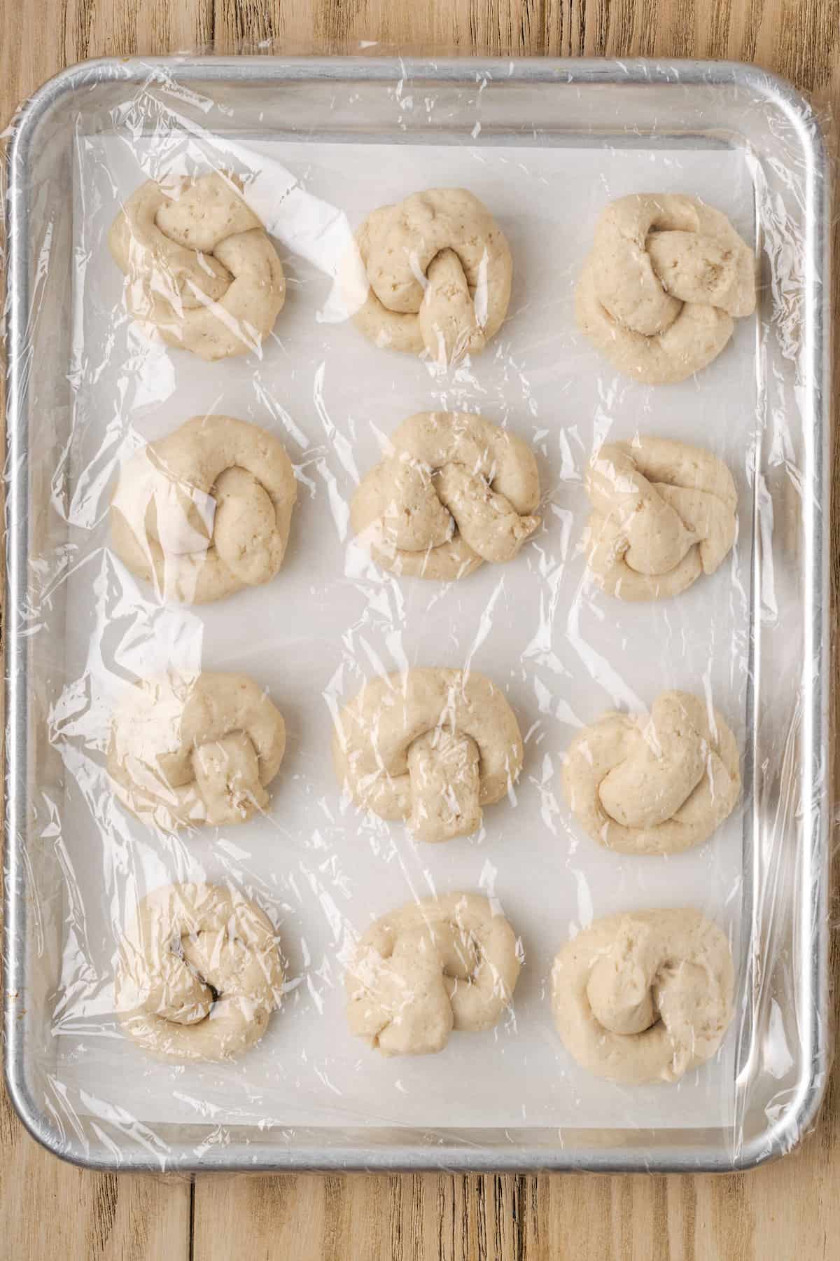 Unbaked gluten-free garlic knots on a lined baking sheet covered in plastic wrap.