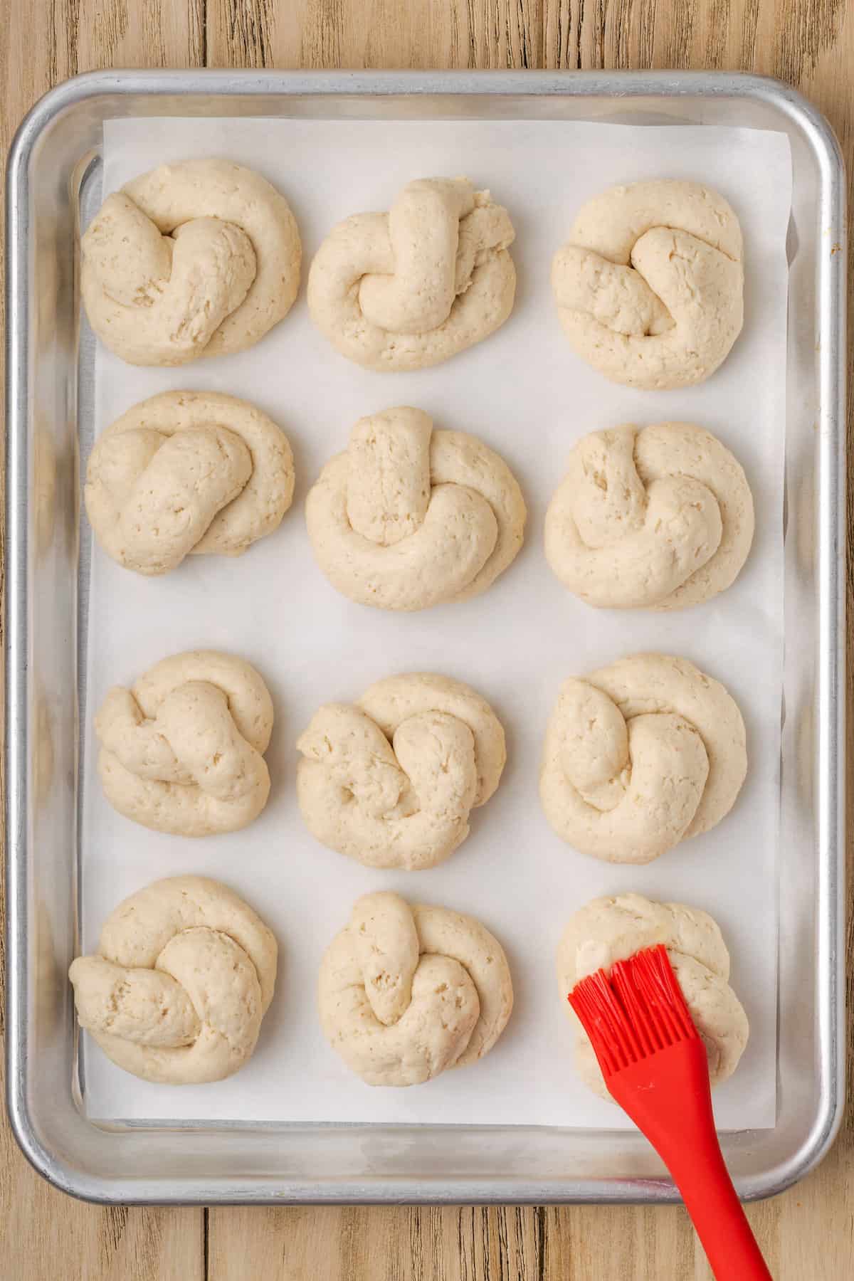 Unbaked gluten-free garlic knots on a baking sheet are brushed with melted butter.
