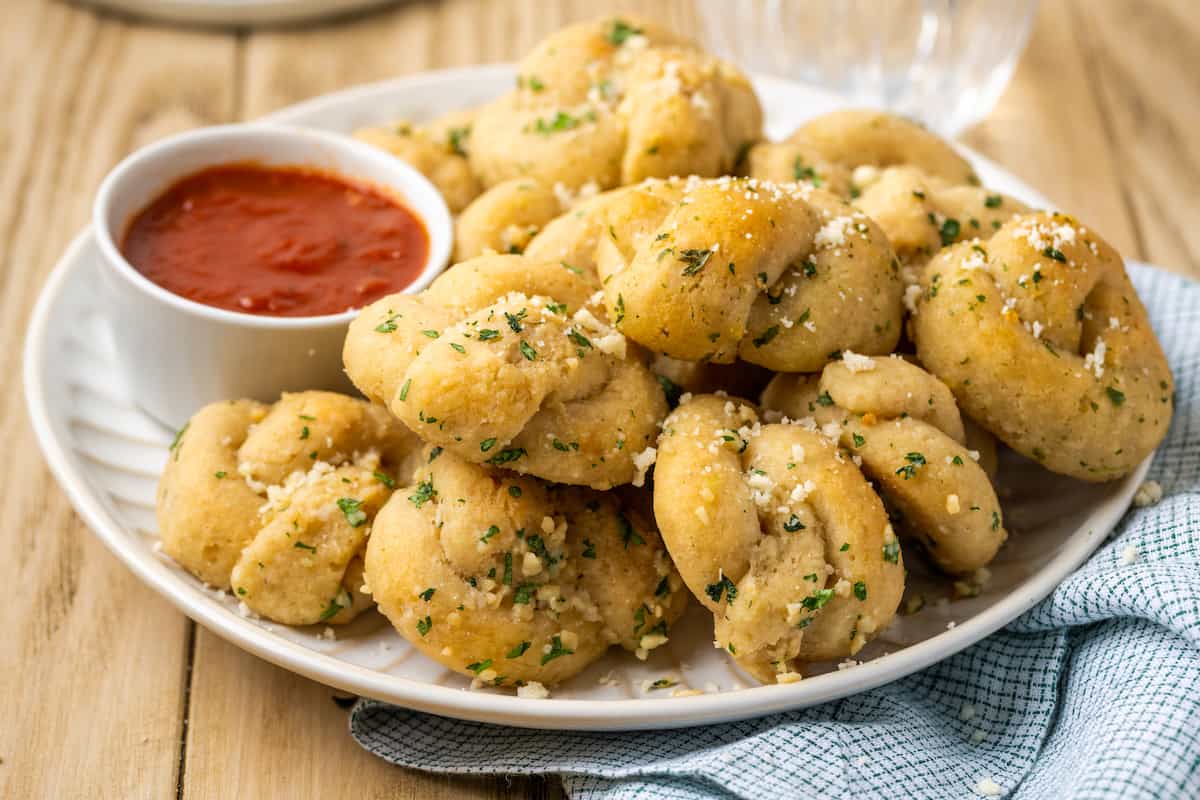 Gluten-free garlic knots stacked on a plate next to a bowl of marinara sauce.
