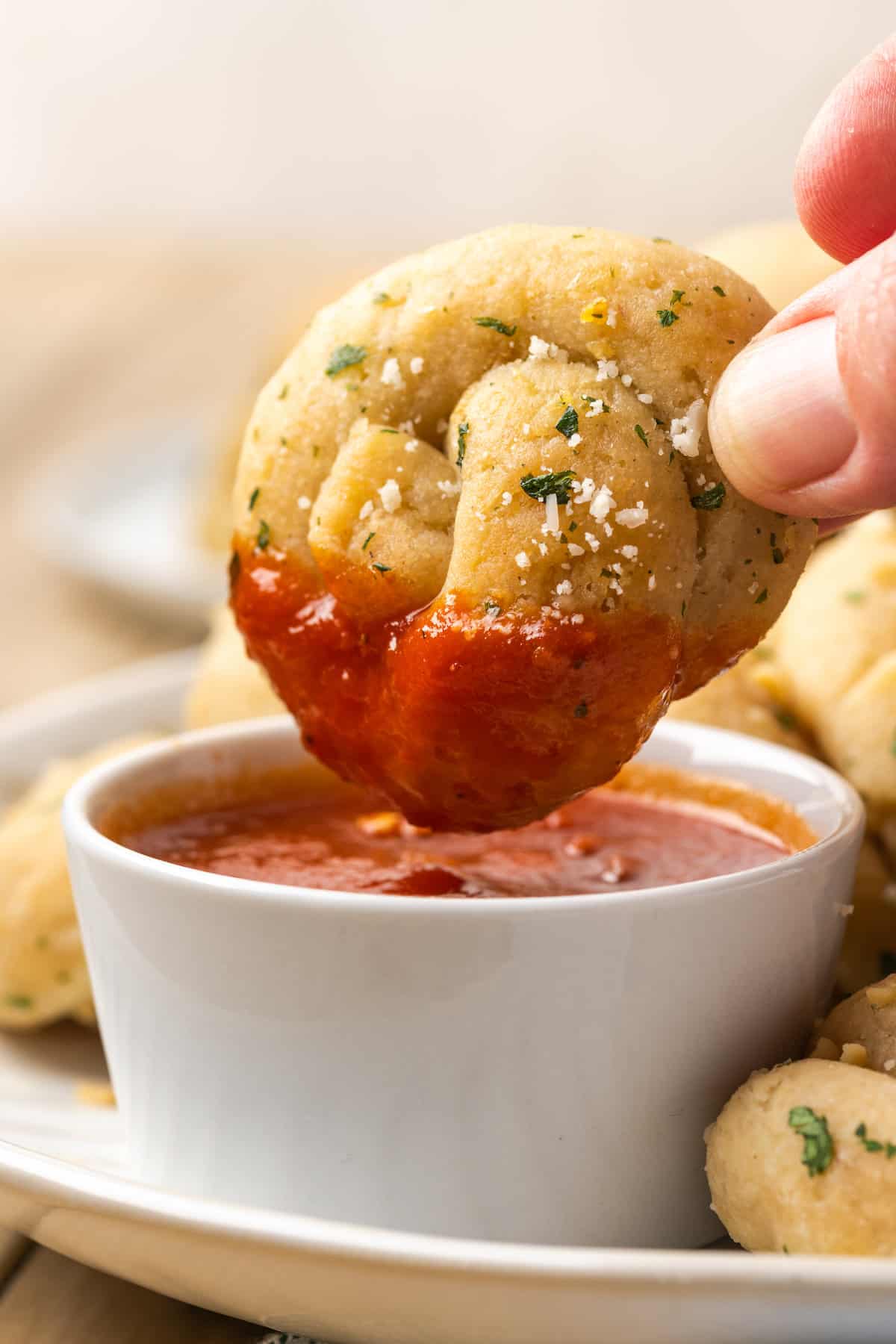 A hand dips a gluten-free garlic knot into a bowl of marinara sauce.