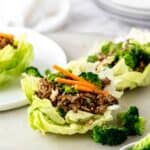 A couple of beef-filled lettuce cups on a serving platter with some small broccoli florets with text overlay that says "Asian Beef Lettuce Wraps".
