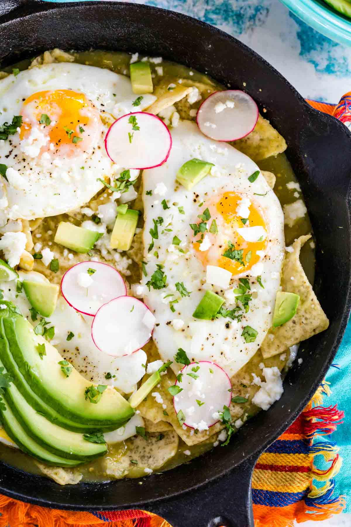 Overhead view of chilaquiles verdes in a cast iron skillet topped with fried eggs, avocado, sliced radishes, and cilantro.