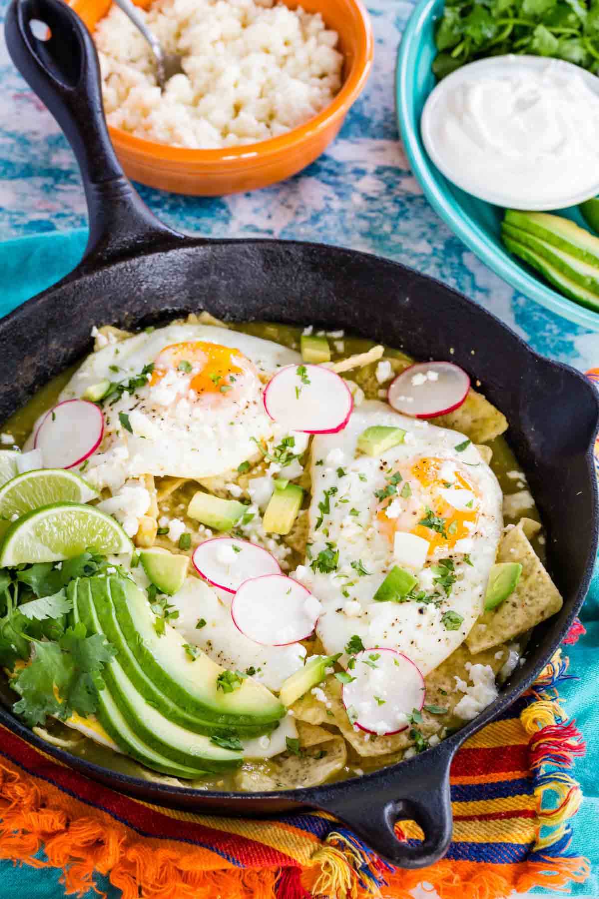 Chilaquiles verdes in a cast iron skillet topped with fried eggs, avocado, sliced radishes, and cilantro.