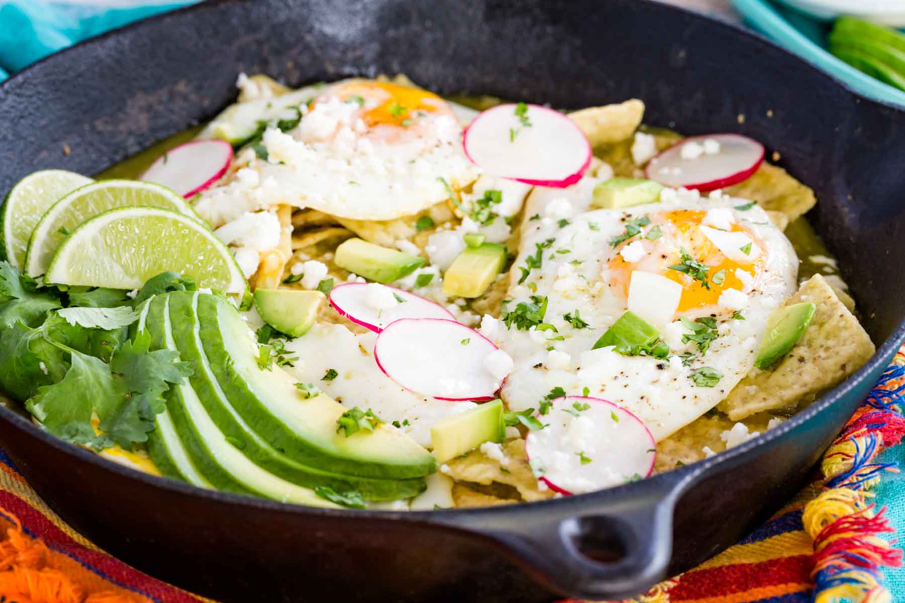 Chilaquiles verdes in a cast iron skillet topped with fried eggs, avocado, sliced radishes, and cilantro.