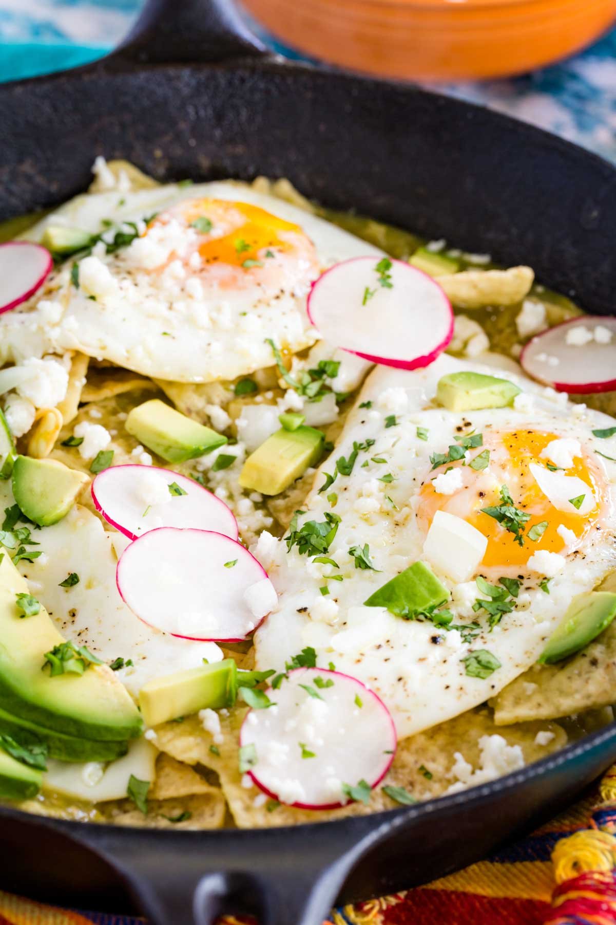 Chilaquiles verdes in a cast iron skillet topped with fried eggs, avocado, sliced radishes, and cilantro.