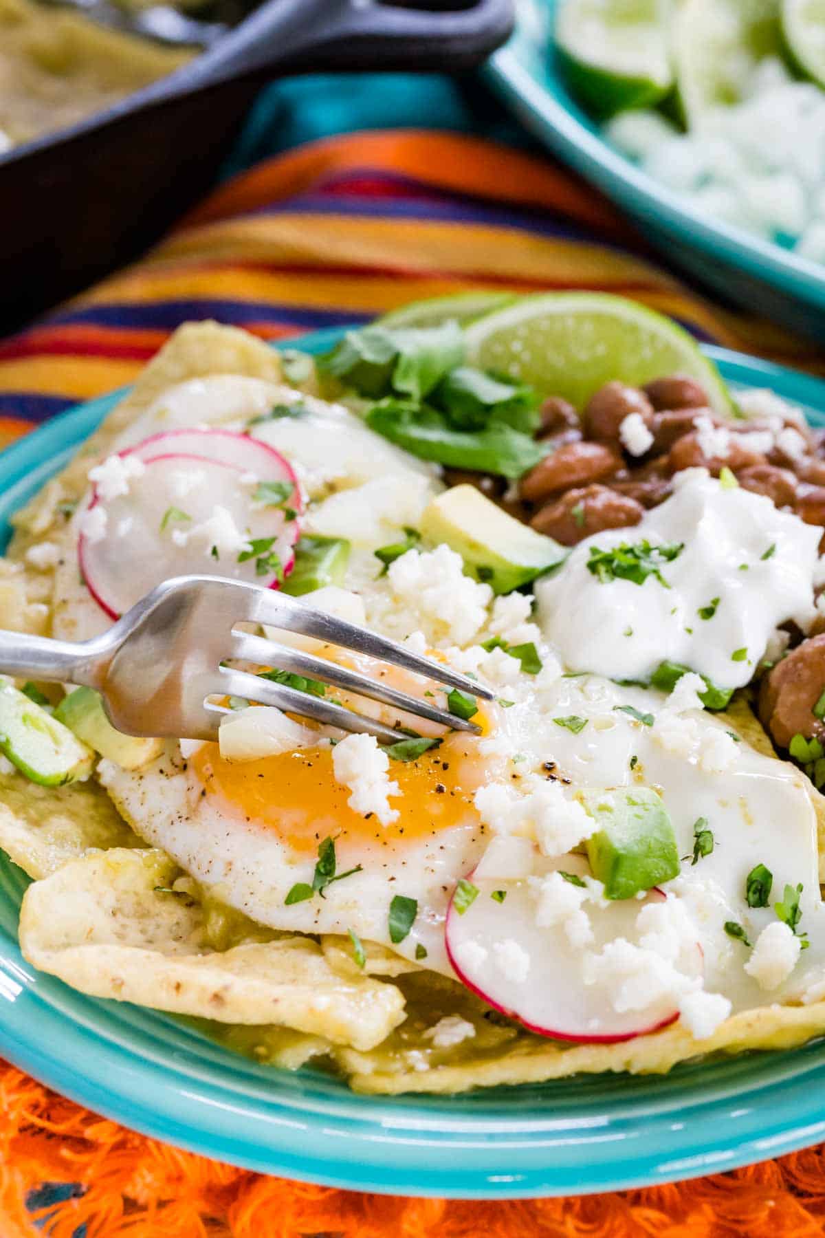 A fork breaks the yolk on a fried egg served on top of chilaquiles verdes topped with garnishes.