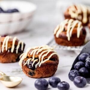 Several flourless blueberry mini muffins with a drizzle on top on a countertop surrounded by a scoop of blueberries.