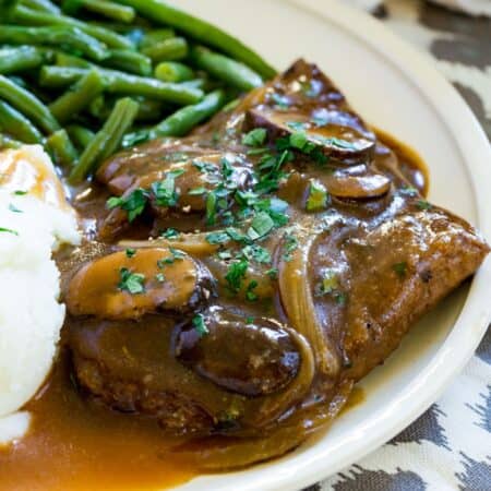A cube steak smothered in gravy with mushrooms and onions on a plate with mashed potatoes and green beans.