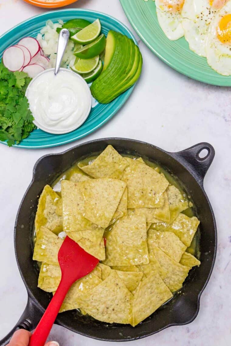 A red spatula stirs tortilla chips in a skillet with salsa verde to coat.
