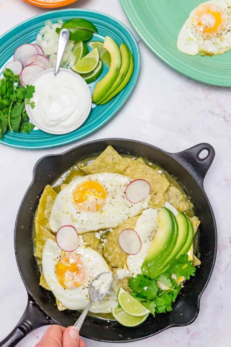 Avocado, radishes, and cheese is added to chilaquiles verdes in a cast iron skillet, next to a plate filled with toppings.