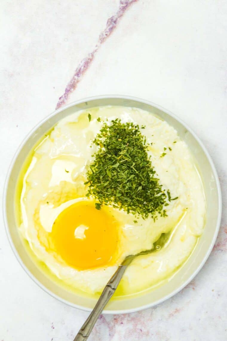 An egg and parsley added to a bowl of ricotta cheese, with a spoon.
