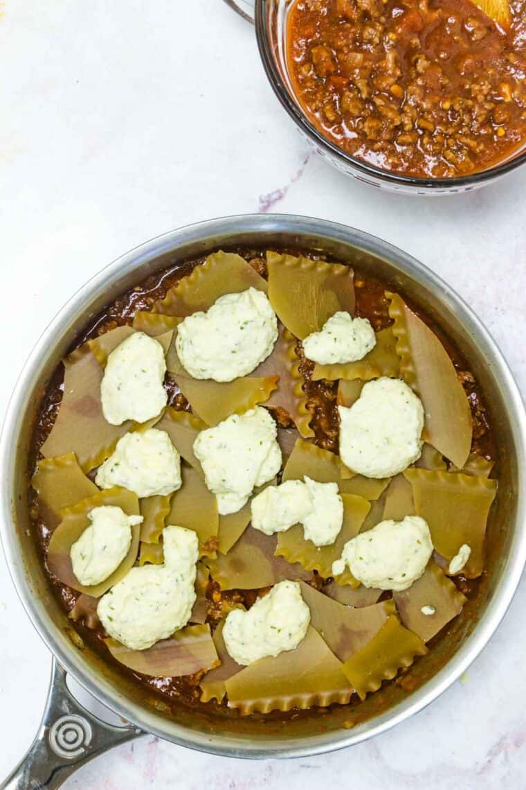 Dollops of ricotta cheese layered over gluten-free lasagna noodles and meat sauce in a skillet.