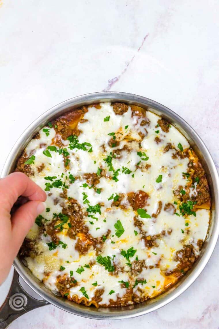 A hand sprinkles parsley over gluten-free lasagna in a skillet.