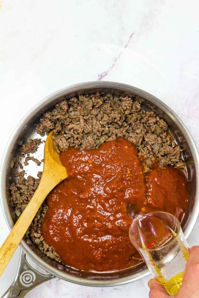 A hand pouring a jar of marinara sauce into ground beef in a skillet.