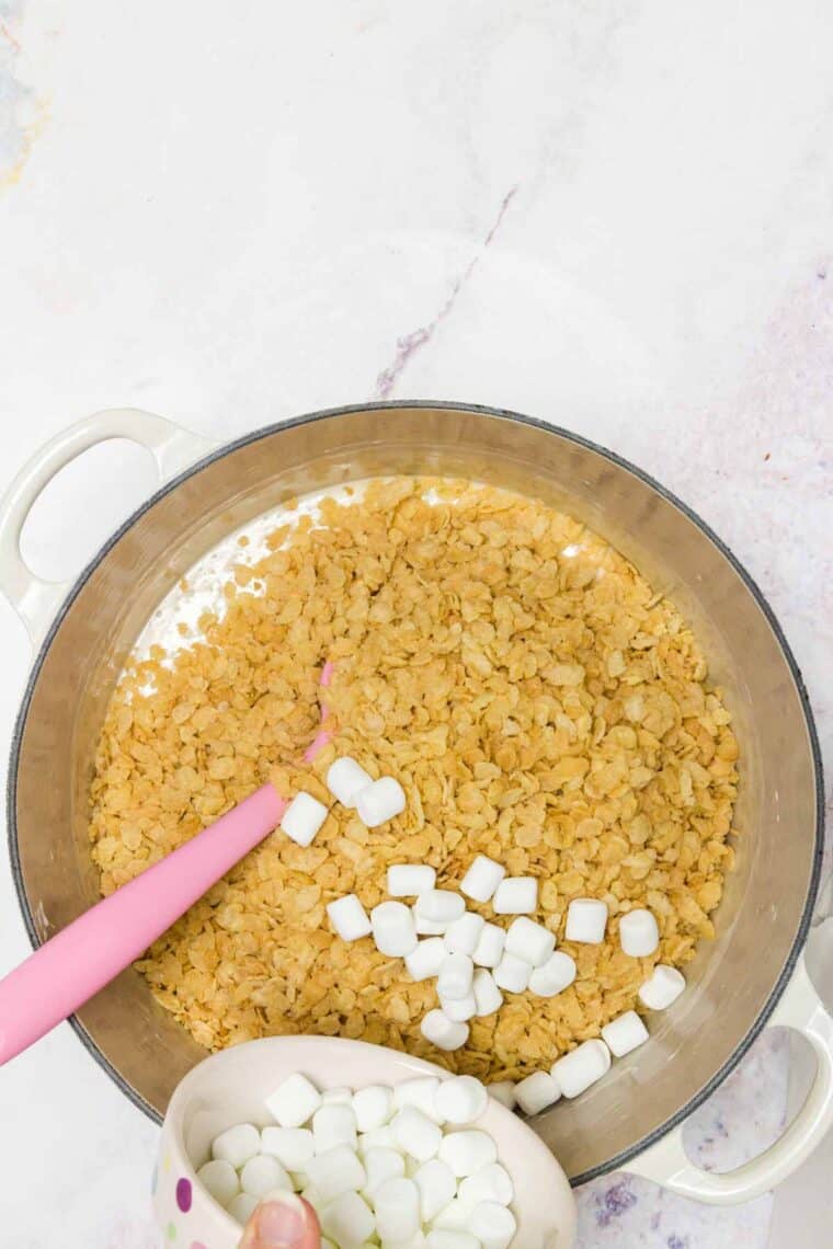 Mini marshmallows are sprinkled into a bowl with melted marshmallows and rice krispies cereal.