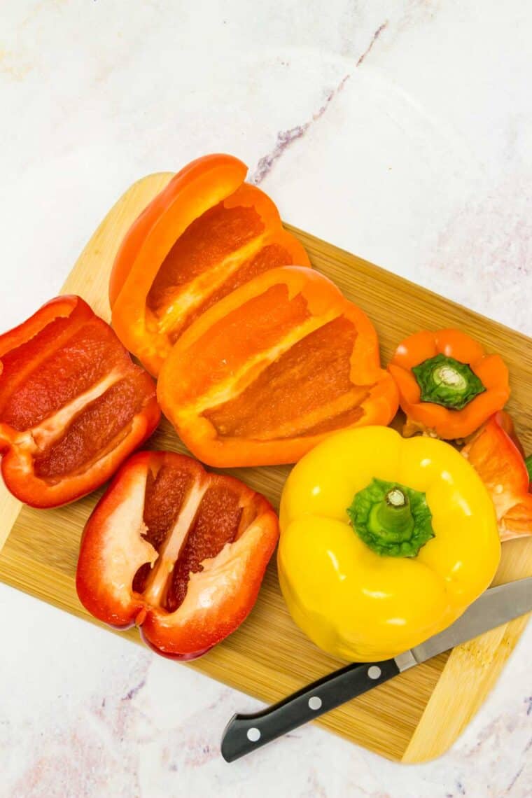 Red, yellow, and orange bell peppers cut in half with their seeds removed on a wooden cutting board.